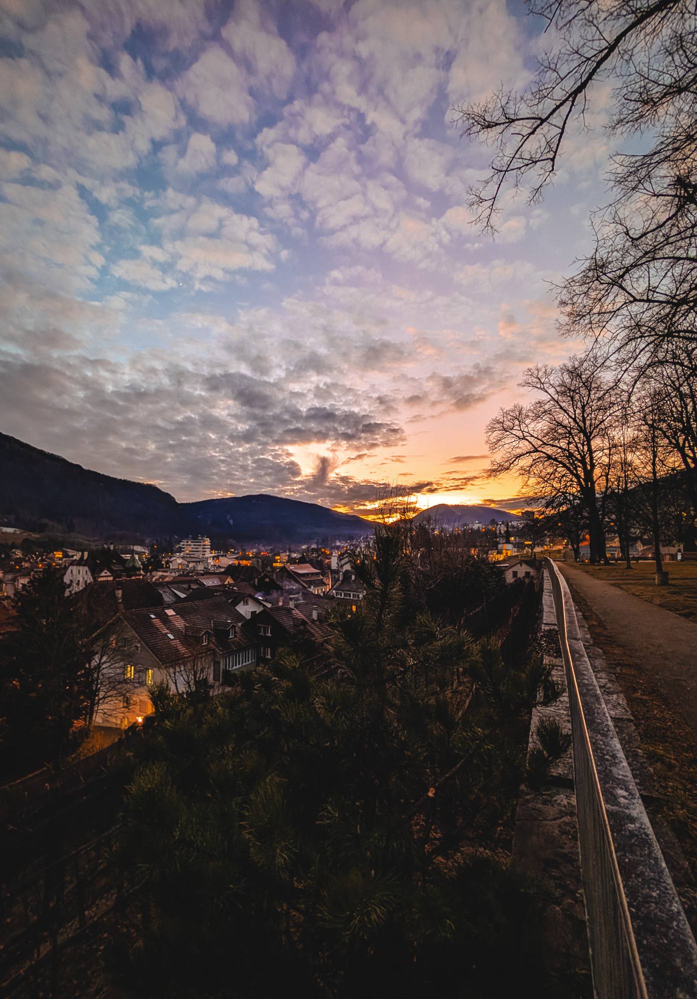 Blick auf eine Stadt bei Sonnenuntergang mit bewölktem Himmel und Bäumen auf einem Weg an einem Hügel.