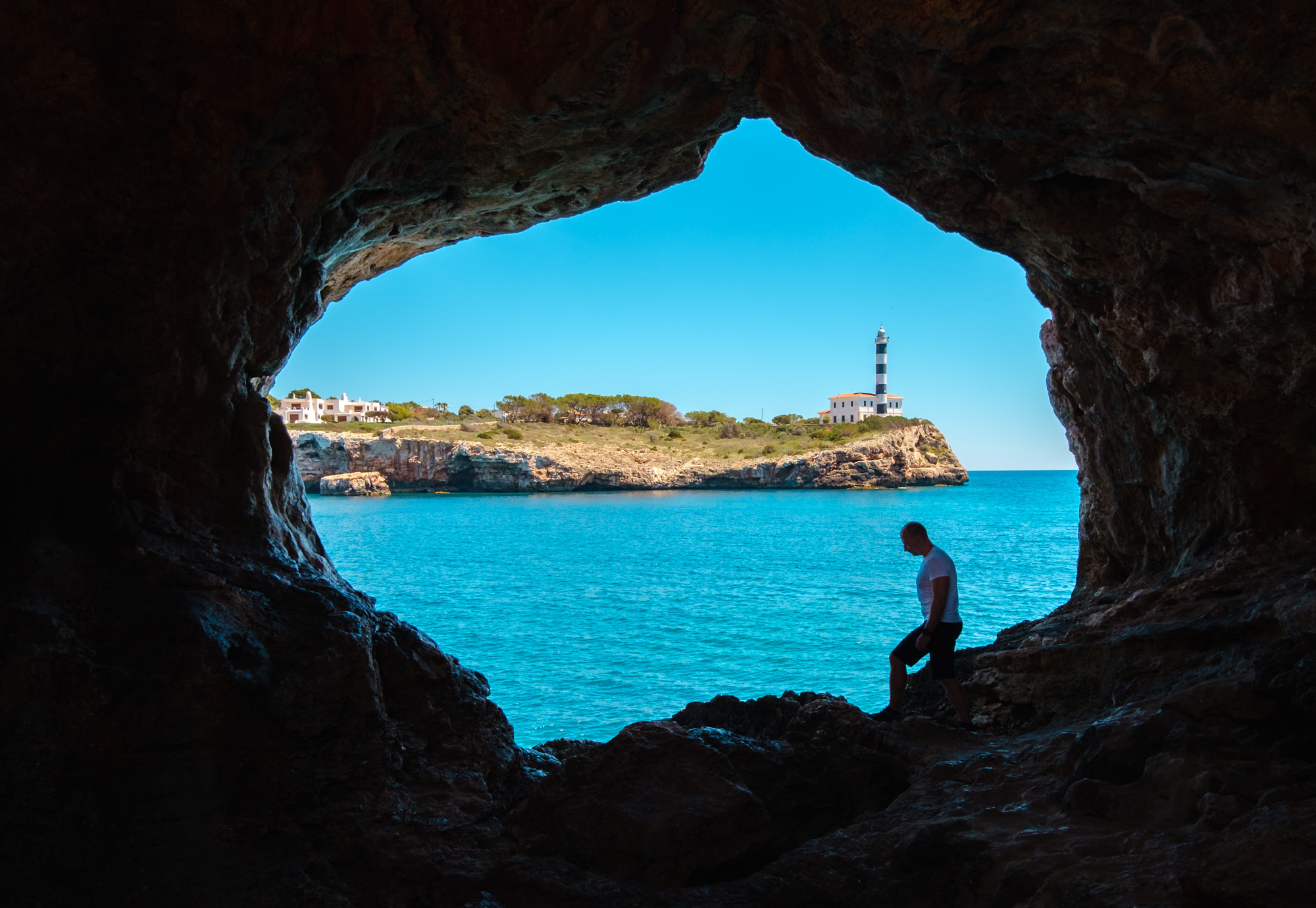 Höhle mit Blick auf Leuchtturm am Meer und einen Mann im Vordergrund, sonniger Tag.