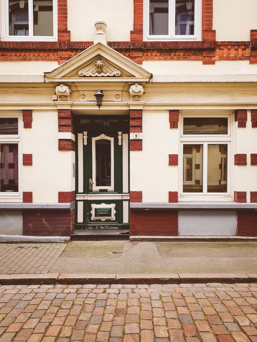 Historische Hausfassade mit grüner Holztür, Fenstern und rotem Backstein auf gepflasterter Straße.