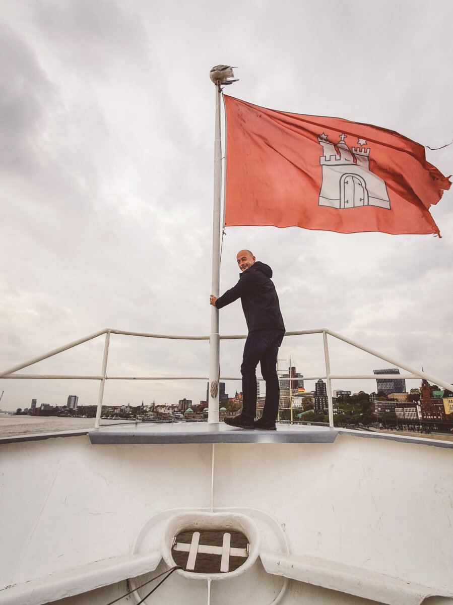 Person steht auf einem Boot und hält die Hamburger Flagge. Im Hintergrund ist die Skyline von Hamburg zu sehen.