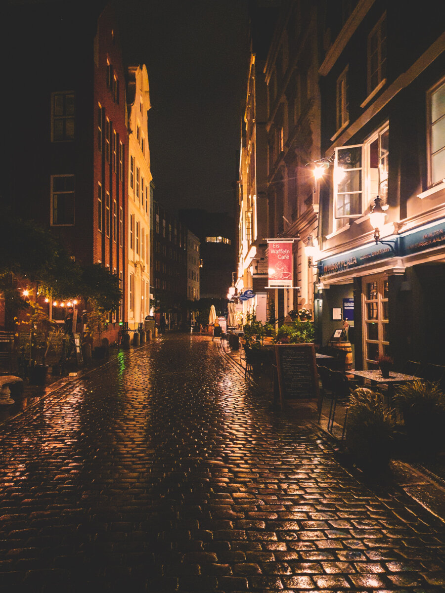 Nasse, gepflasterte Gasse bei Nacht mit beleuchteten Gebäuden und Cafés auf beiden Seiten, unter dunklem Himmel.