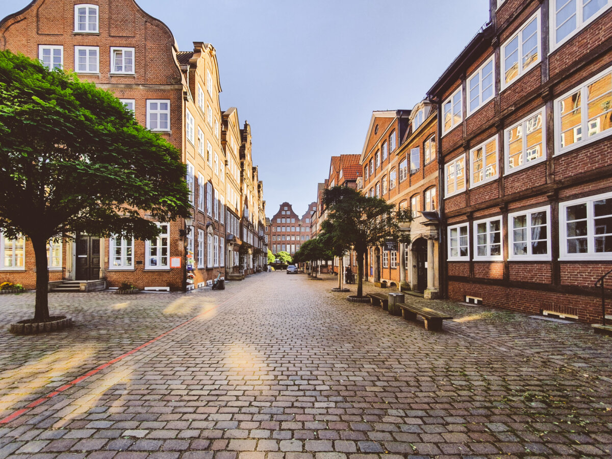 Pflasterstraße mit historischen Backsteinhäusern und Bäumen bei sonnigem Wetter.