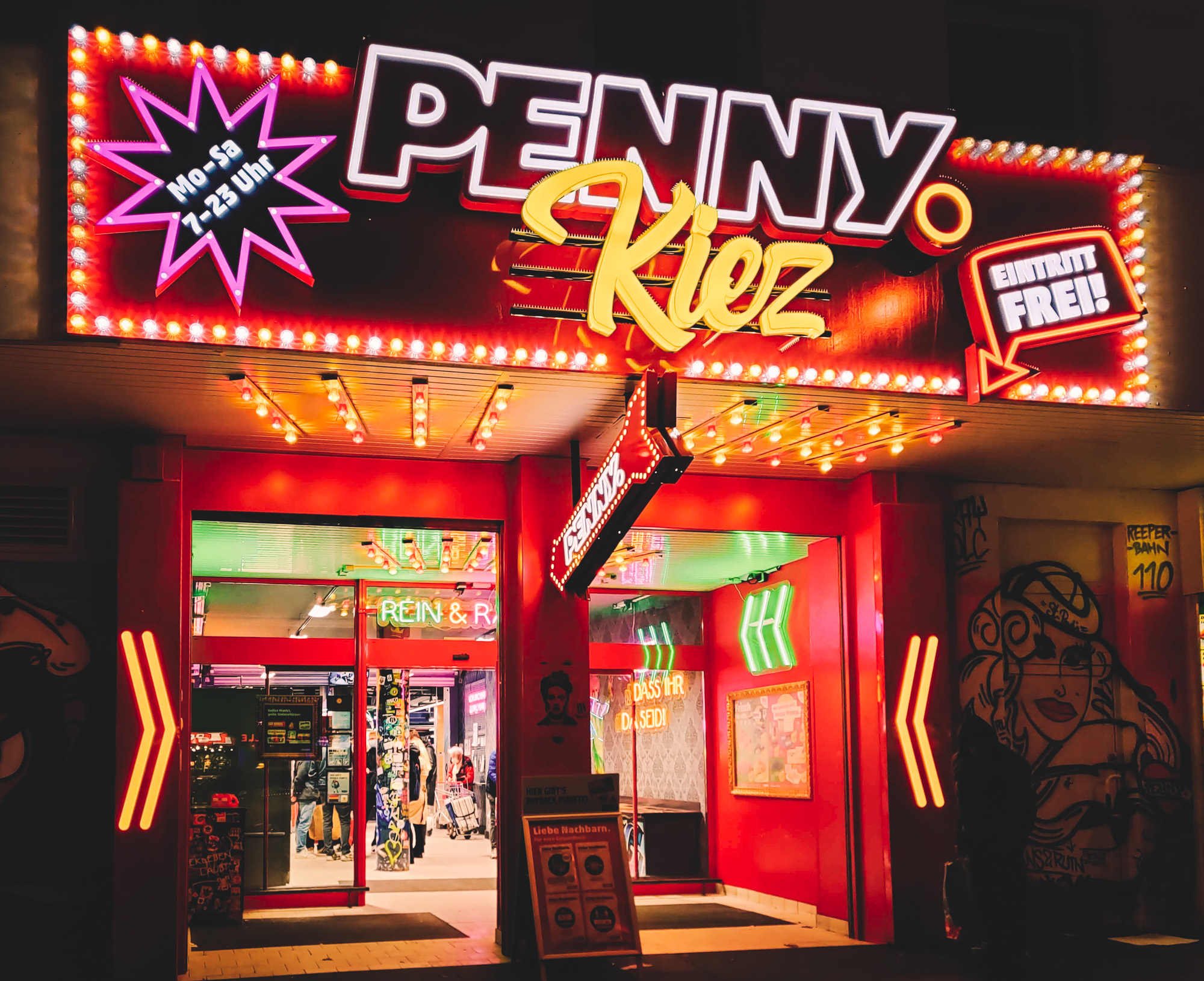 Neonbeschilderter Eintritt eines Penny-Markts in Hamburg bei Nacht. Bunte Lichter und lebhafte Atmosphäre.