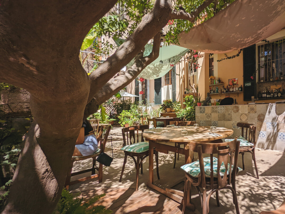 Sonniger, gemütlicher Restaurantgarten mit Baum, Holztisch, Stühlen und einer Bar im Hintergrund.