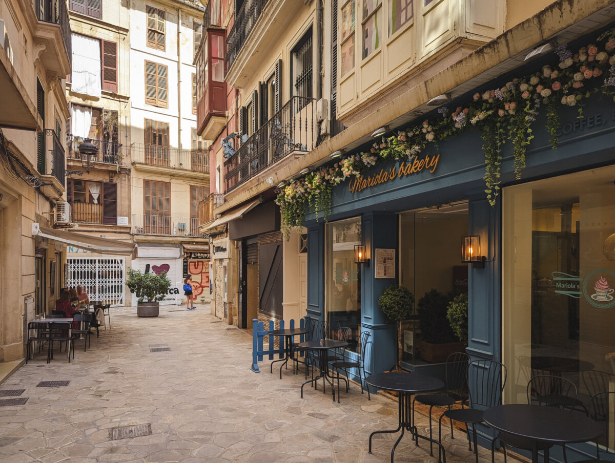 Malersche Gasse mit Außensitzplätzen von Mariola's Bakery, gerahmt von historischen Gebäuden, sonniger Tag.