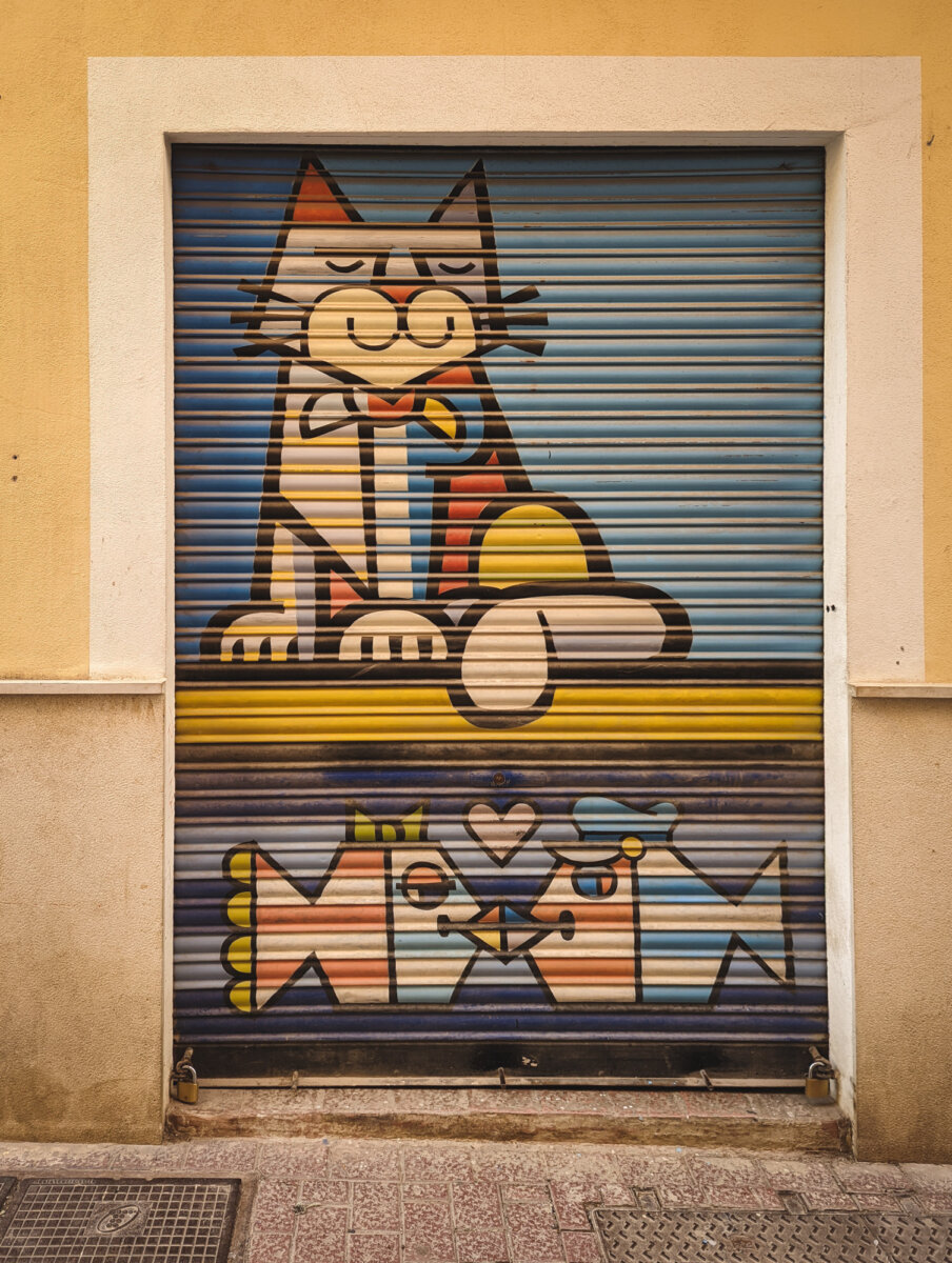 Buntes Graffiti eines lächelnden Hundes und zwei Fischen auf einem Rolltor an gelber Hauswand.