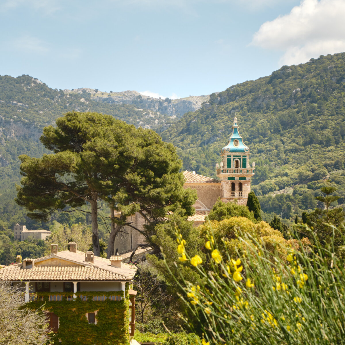 Turm mit grünlich-blauem Dach in gebirgiger Landschaft, umgeben von Bäumen und Blumen unter blauem Himmel.