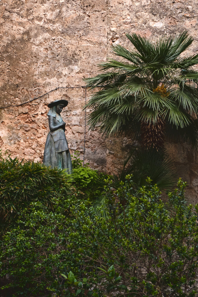 Bronzestatue einer Frau im Hut, umgeben von üppigem Grün und einer Steinmauer im Hintergrund.