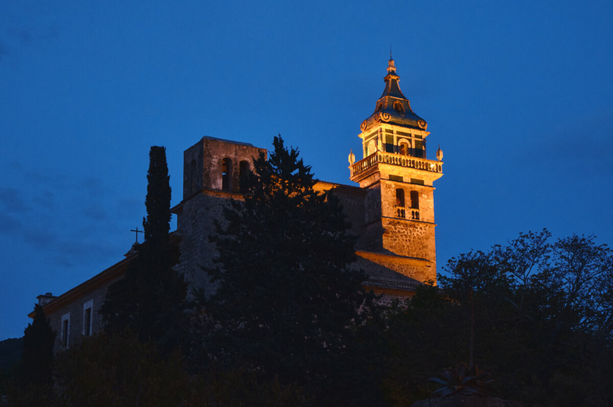 Beleuchteter Glockenturm eines Klosters bei Abenddämmerung, umgeben von Bäumen und klarem Abendhimmel.
