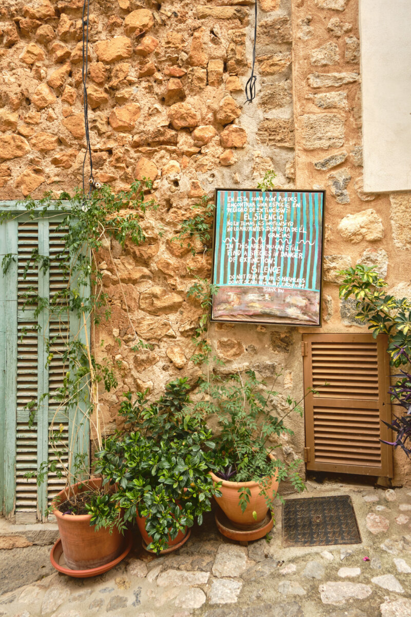 Steinwand mit spanischem Schild, Kletterpflanzen in Töpfen und geschlossenen Fensterläden in einer Gasse.