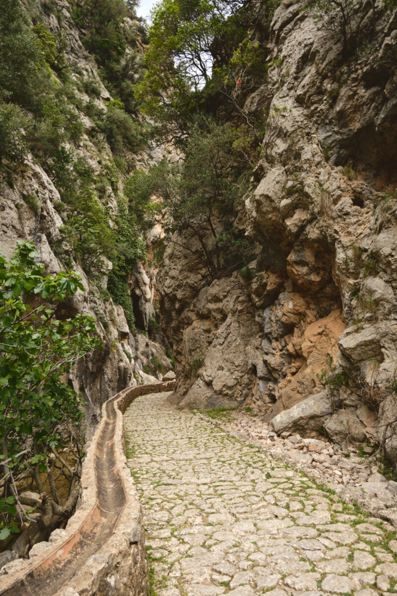 Steinweg durch eine schmale Schlucht mit hohen, bewachsenen Felsen und einem kleinen Wasserkanal rechts des Weges.