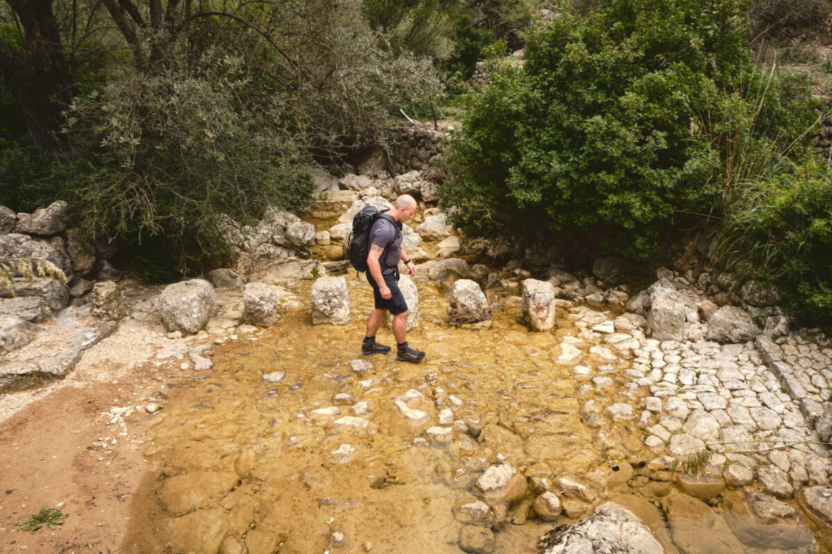 Wanderer mit Rucksack überquert einen flachen, steinigen Fluss in einer bewaldeten, felsigen Landschaft.