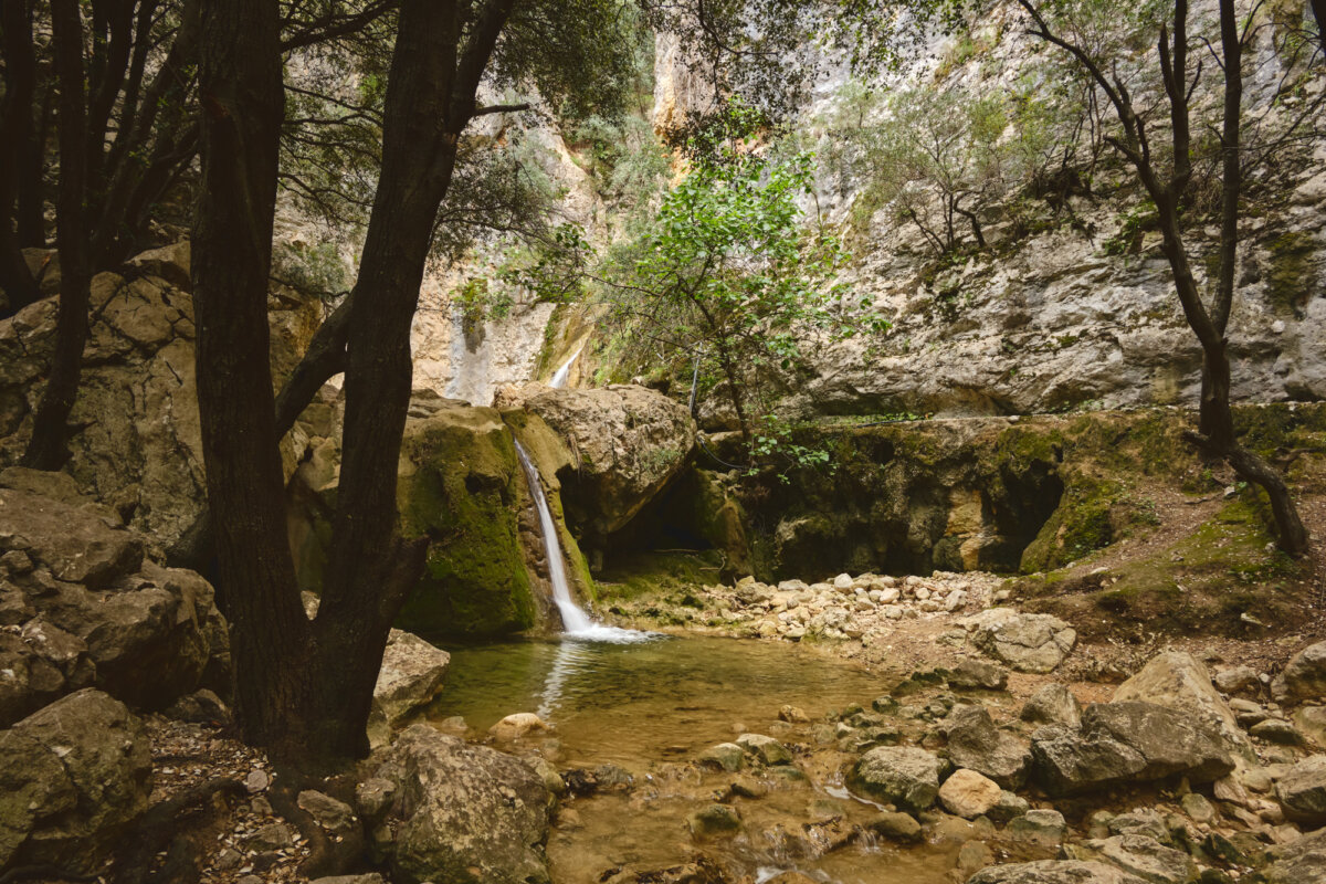 Wasserfall in einem bewaldeten Gebirgstal mit Felsen und klarem Wasserbecken im Vordergrund.