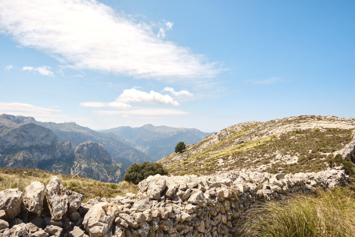 Steinmauer in mediterraner Berglandschaft unter blauem Himmel mit vereinzelten Wolken.