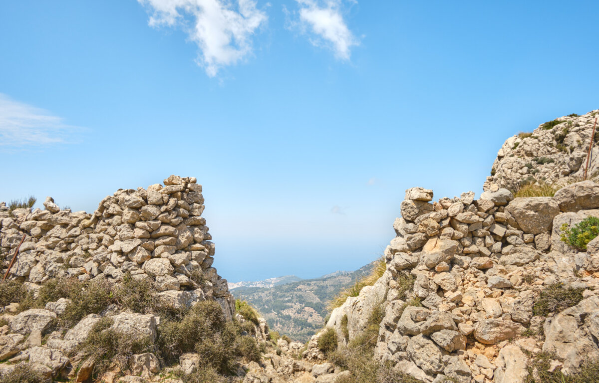 Steinmauer öffnet den Blick auf das Meer und die Berge unter klarem Himmel in der Ferne.
