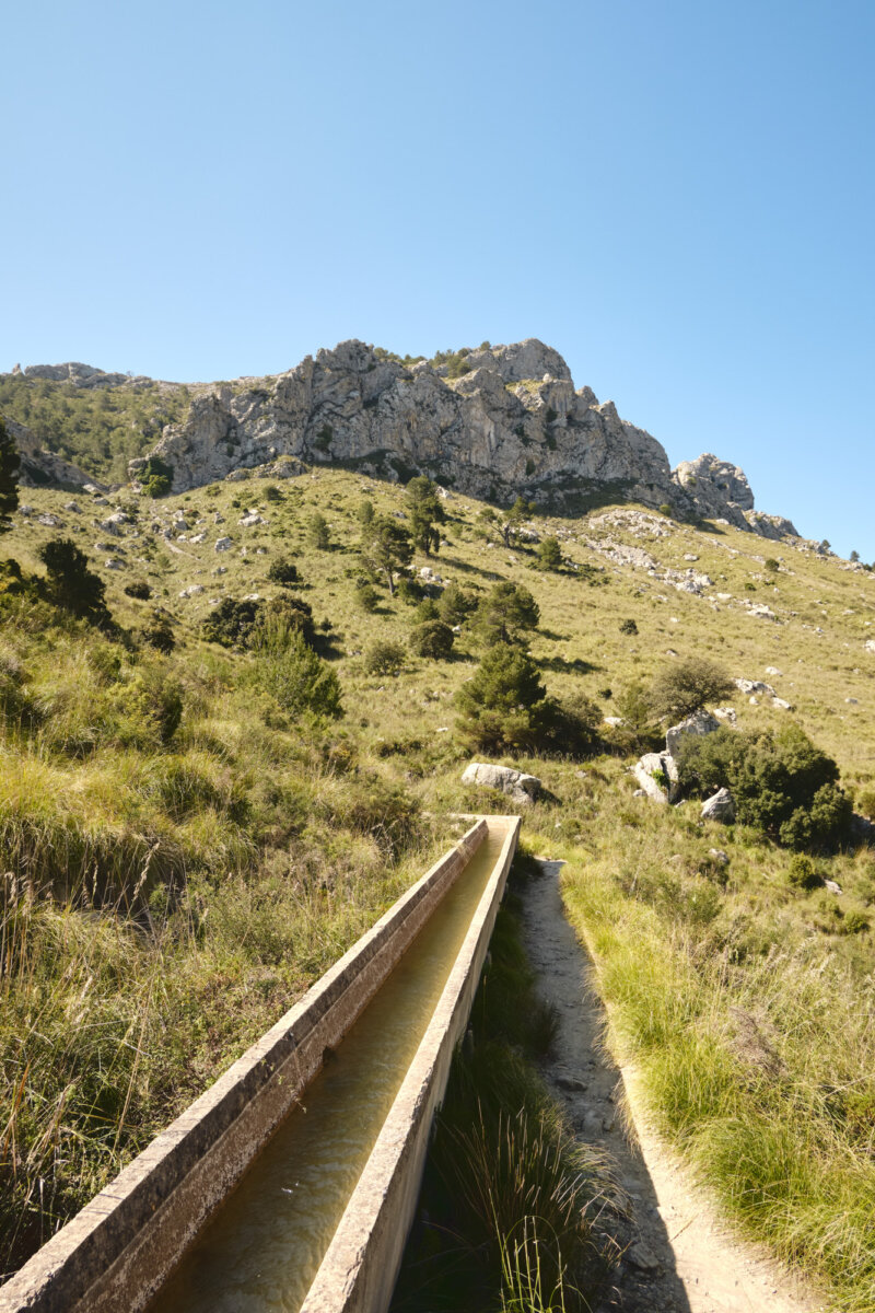 Langer Bewässerungskanal führt durch grüne Landschaft mit felsigem Hügel bei klarem Himmel im Hintergrund.