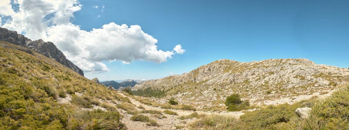 Weite Berglandschaft mit felsigen Hügeln, grünem Gras und blauem Himmel mit großen weißen Wolken.