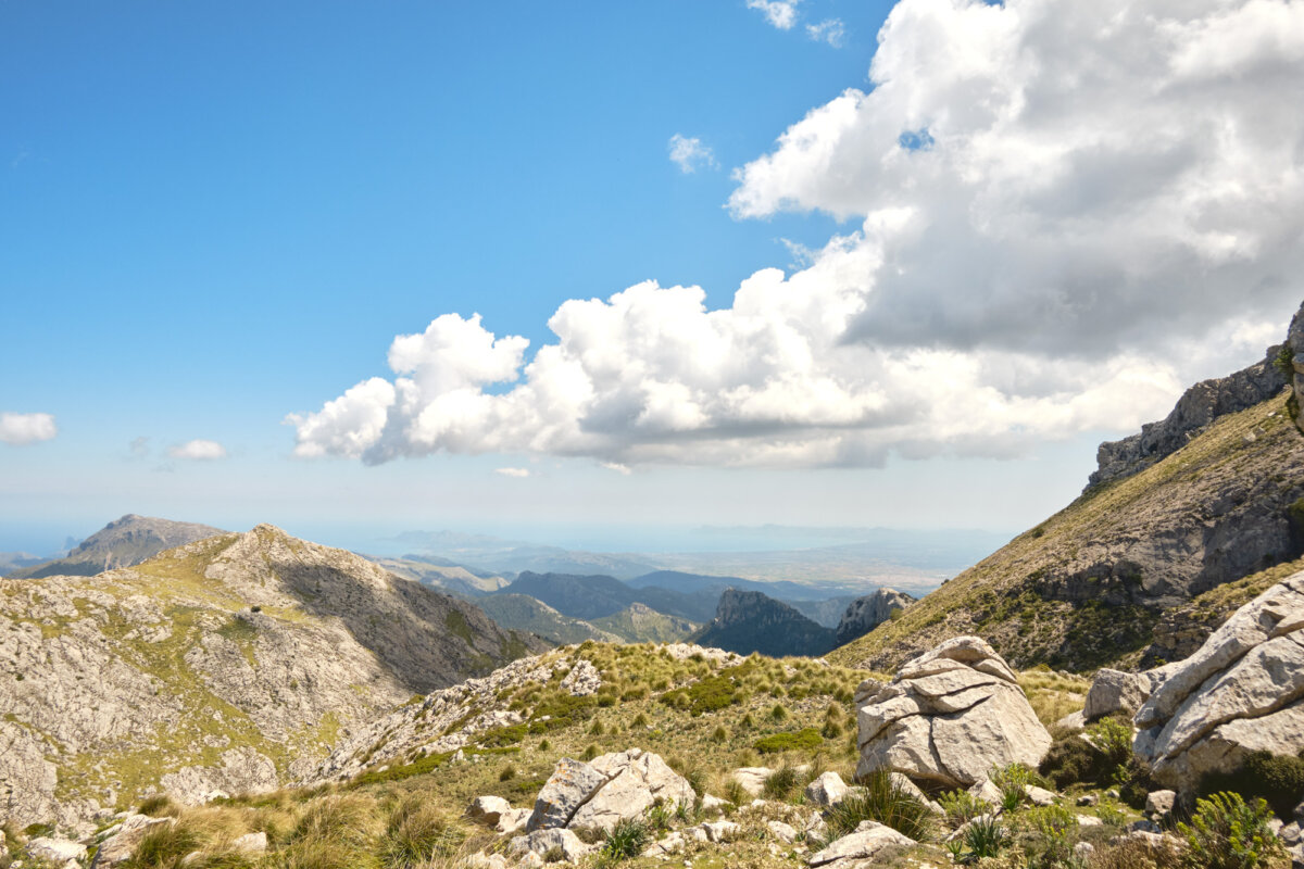Weitläufige Berglandschaft unter blauem Himmel mit weißen Wolken, felsige Hügel und Gräser im Vordergrund.