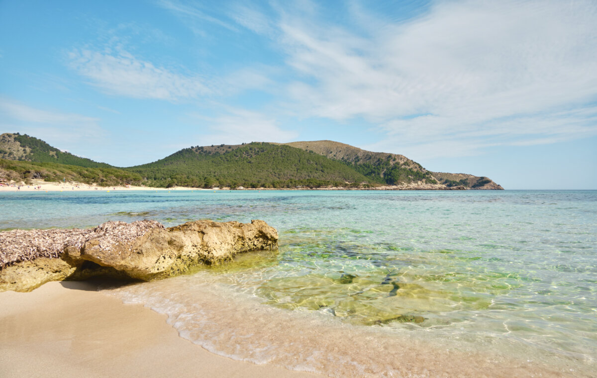 Klares Wasser und sandiger Strand mit grünen Hügeln im Hintergrund unter blauem Himmel an einem sonnigen Tag.