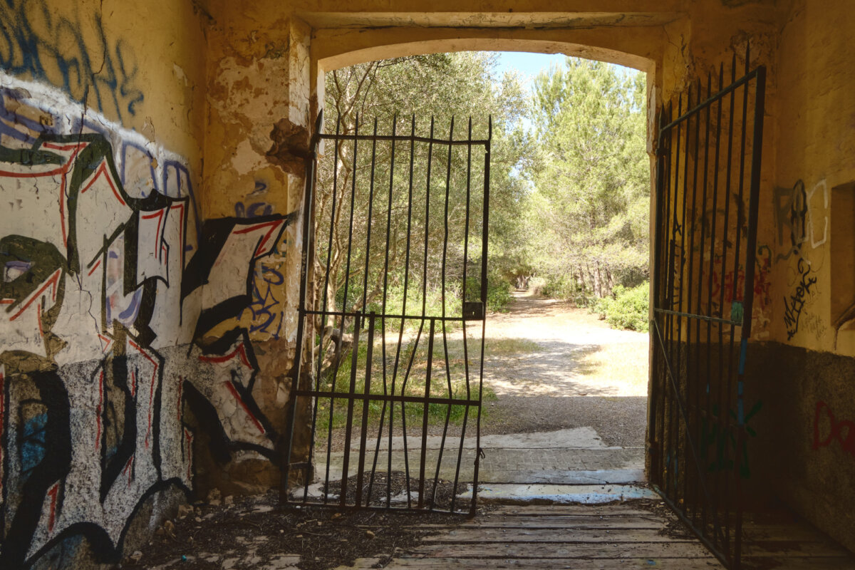Verfallenes Tor mit Graffiti, das zu einem sonnigen, von Bäumen gesäumten Pfad führt.
