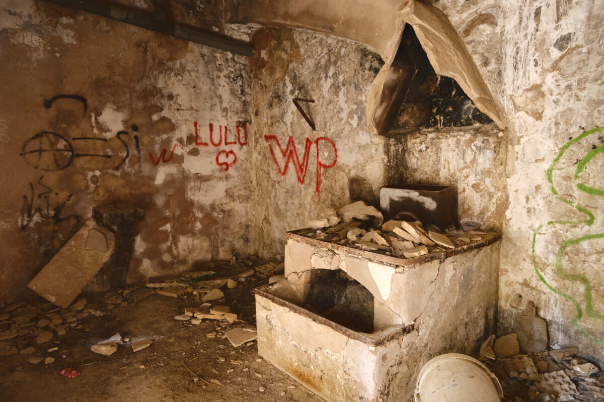 Verfallener Raum mit Graffiti und kaputtem Ofen. Wände und Boden sind mit Schutt bedeckt.