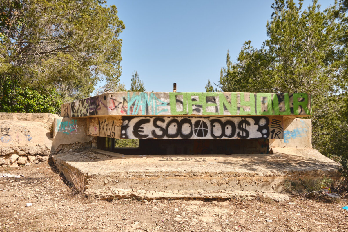 Betonbunker im Wald mit bunten Graffitis, darunter das Zeichen für Anarchie, Europäisches Euro- und US-Dollar-Symbol.