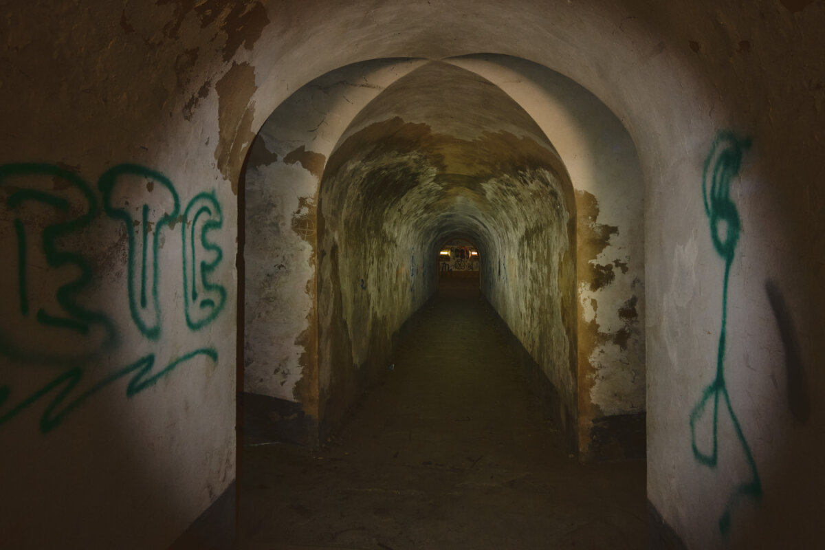 Dunkler, gewölbter Tunnel mit Graffiti an den Wänden, der in die Ferne führt. Schummriges Licht beleuchtet den Weg.