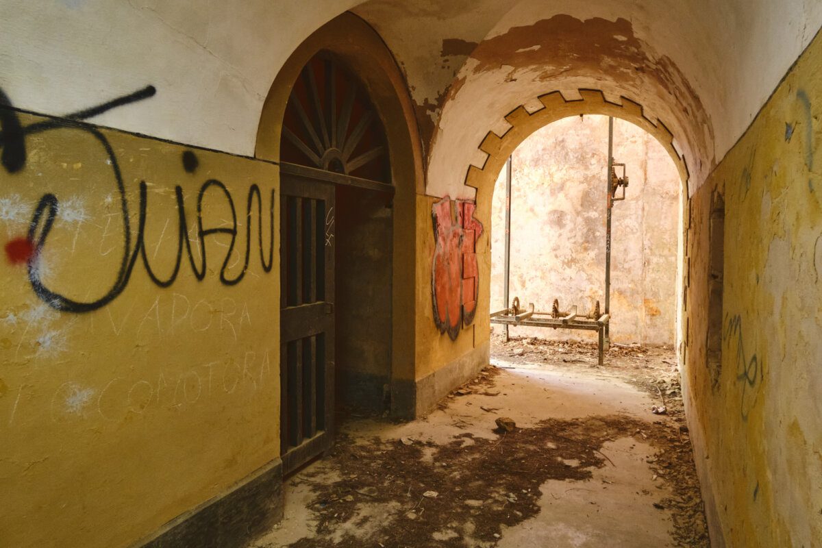 Verlassener, bogenförmiger Gang mit Graffiti an den Wänden und bröckelndem Putz, ein Torbogen im Hintergrund.