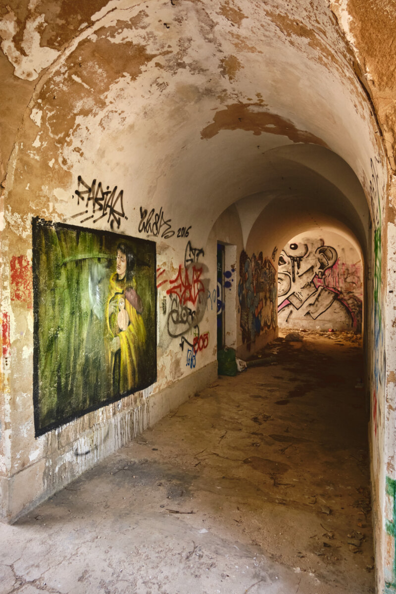 Verlasstener Tunnel mit Graffiti-Kunst an den Wänden, darunter ein Gemälde einer Person in gelbem Umhang.