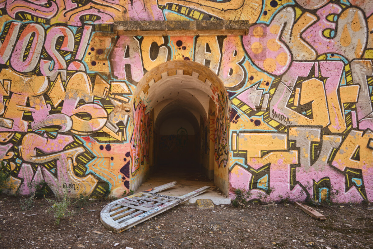 Verlassener Tunnel mit buntem Graffiti und kaputtem Holztor auf erdigem Boden. Abenteuerliche Atmosphäre.