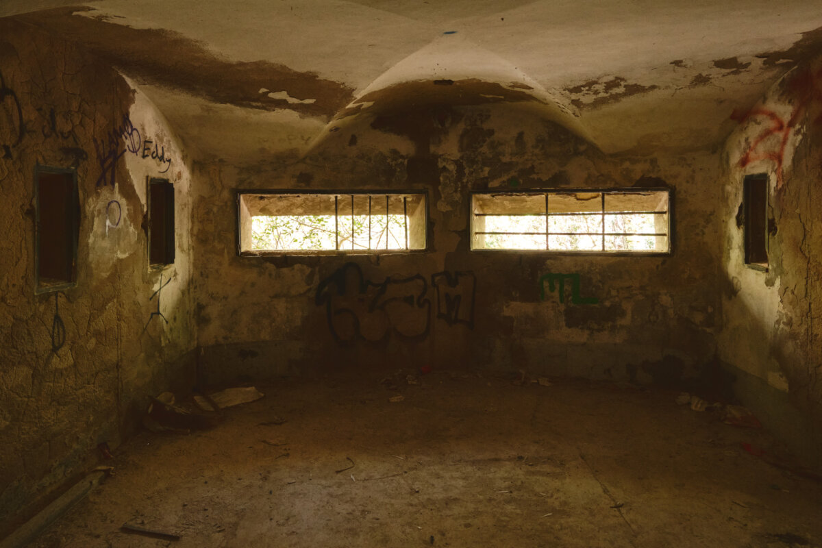 Verlassener Raum mit grauen Wänden und Graffiti, durch vergitterte Fenster fällt Licht ein.