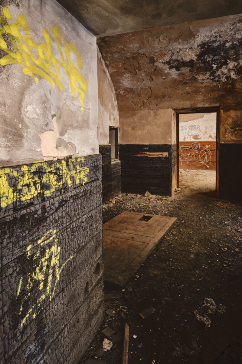 Verfallener Raum mit Graffiti an den Wänden, bröckelndem Putz und Schutt auf dem Boden. Mysteriöse, verlassene Atmosphäre.