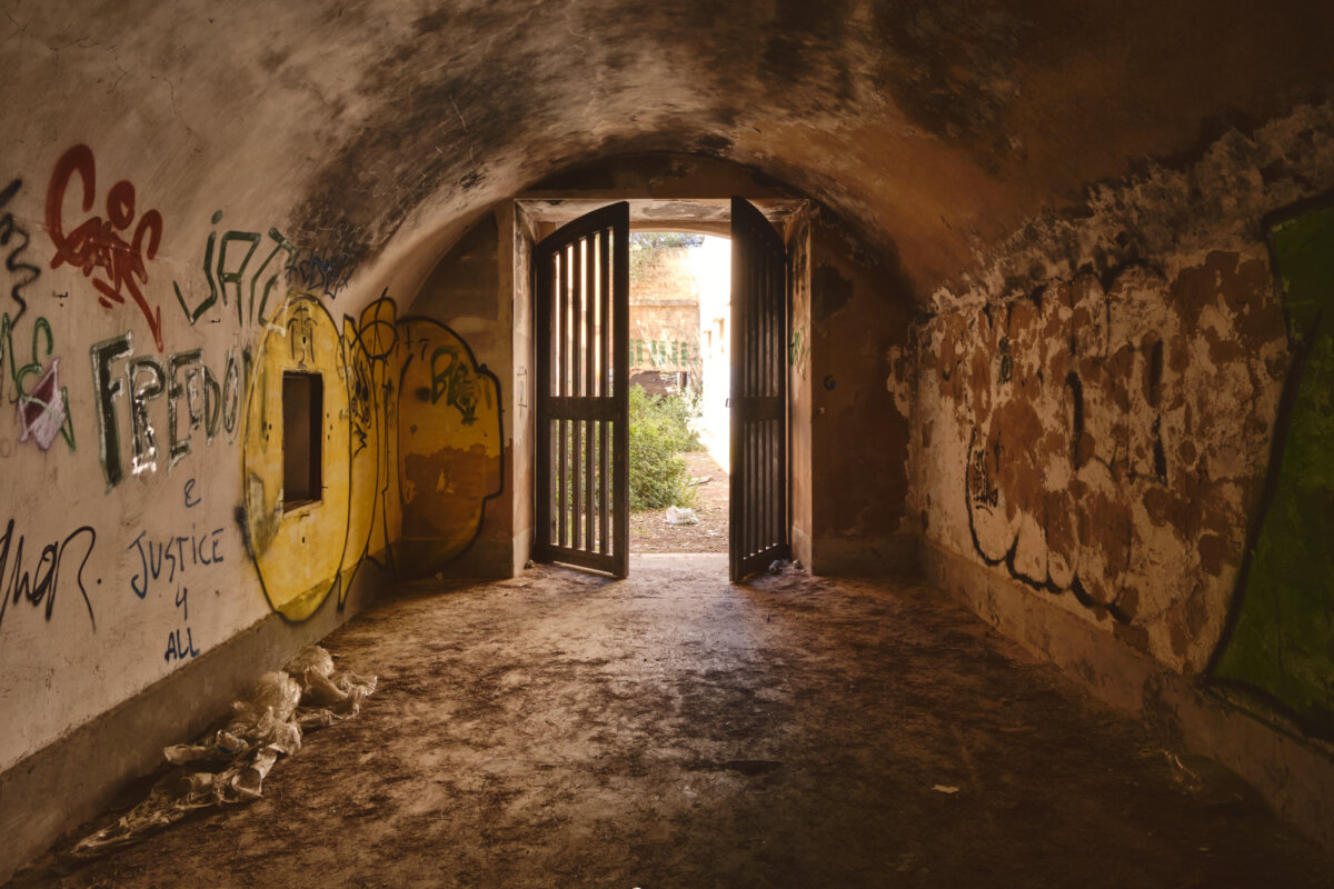 Verlassener Tunnel mit Graffiti an den Wänden, eine geöffnete Holztür führt ins Freie, Tageslicht fällt ein.