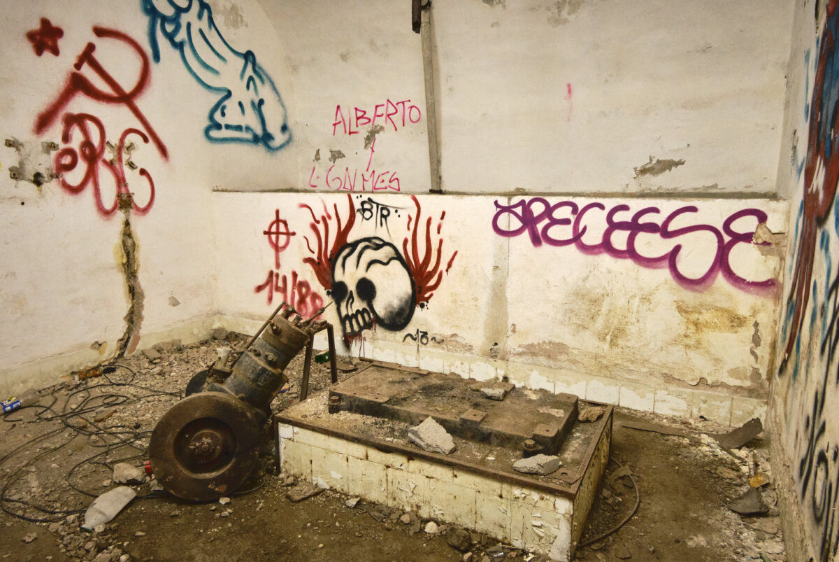 Verlassener Raum mit Graffiti an den Wänden, darunter ein Totenkopf und Schriftzüge. Boden ist mit Schutt bedeckt.