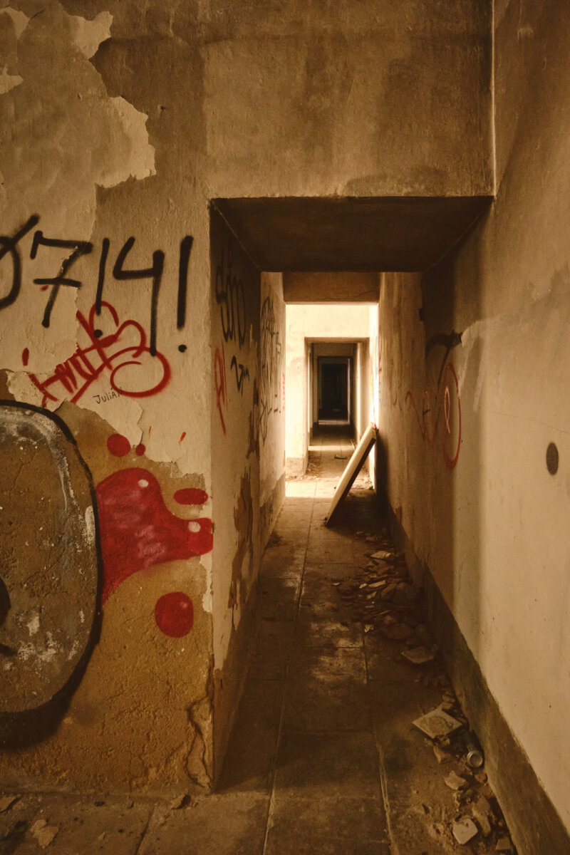 Verlassener, mit Graffiti bedeckter Flur, der in Räume mit schwachem Licht und umgestürzter Tür führt.