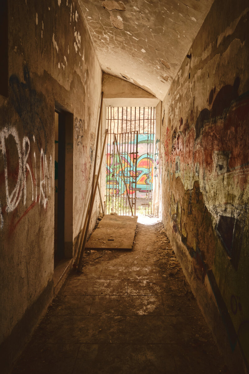 Verlassener, graffitibedeckter Korridor mit Gittern. Licht fällt durch das Tor, Betonboden voller Schmutz und Trümmer.