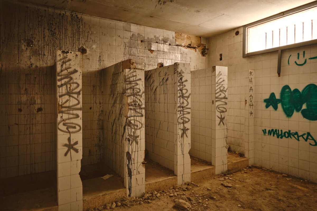 Verlassenes Badezimmer mit Graffiti an den Wänden, bröckelnde Fliesen und Schmutz bedecken den Boden.