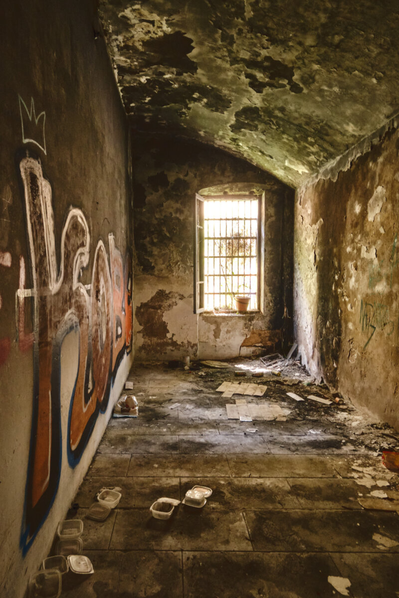 Verlassener Raum mit Graffiti an der Wand, abblätternder Putz und vergittertes Fenster, schummriges Licht.