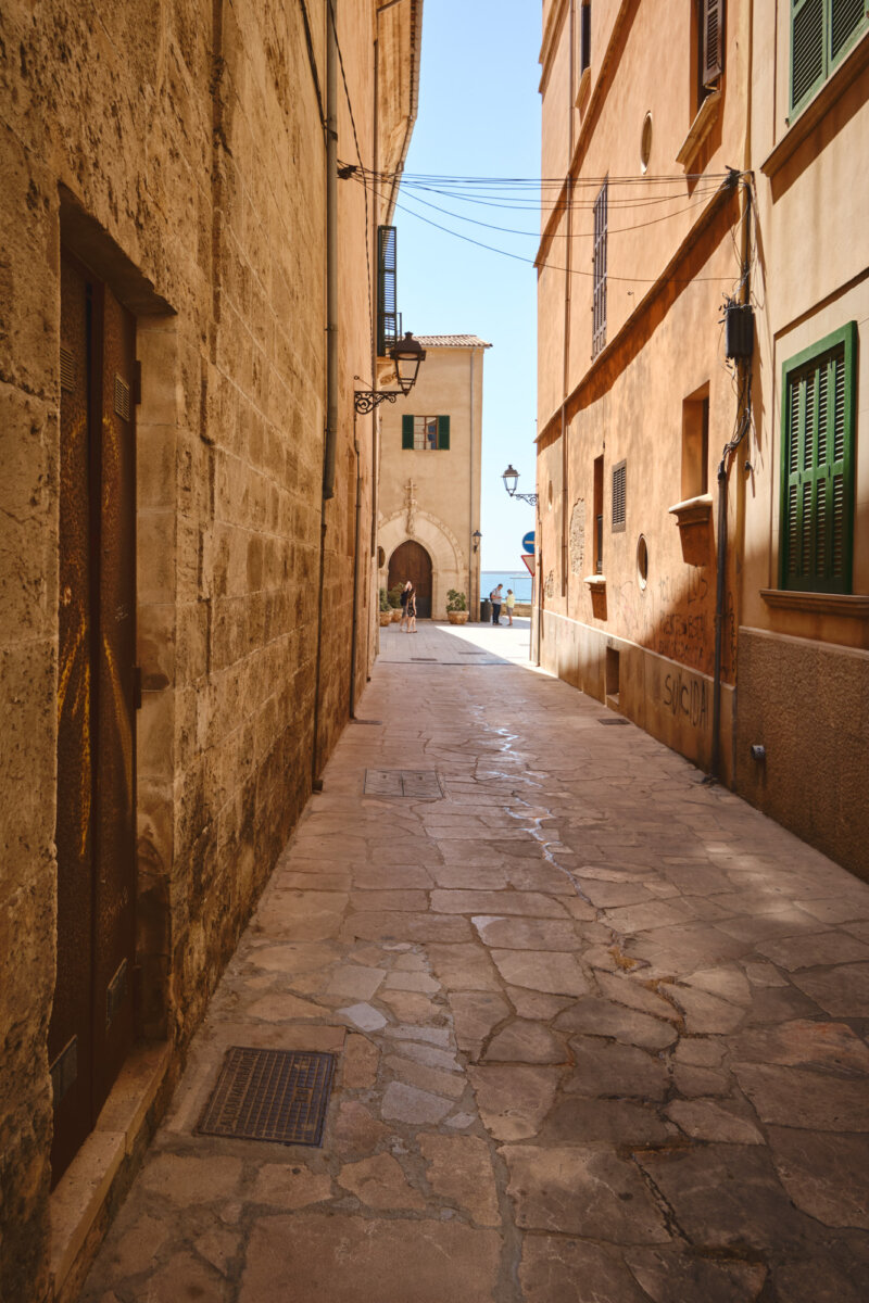 Eine schmale Gasse mit alten Gebäuden in der Altstadt bei hellem Sonnenlicht, im Hintergrund das Meer sichtbar.