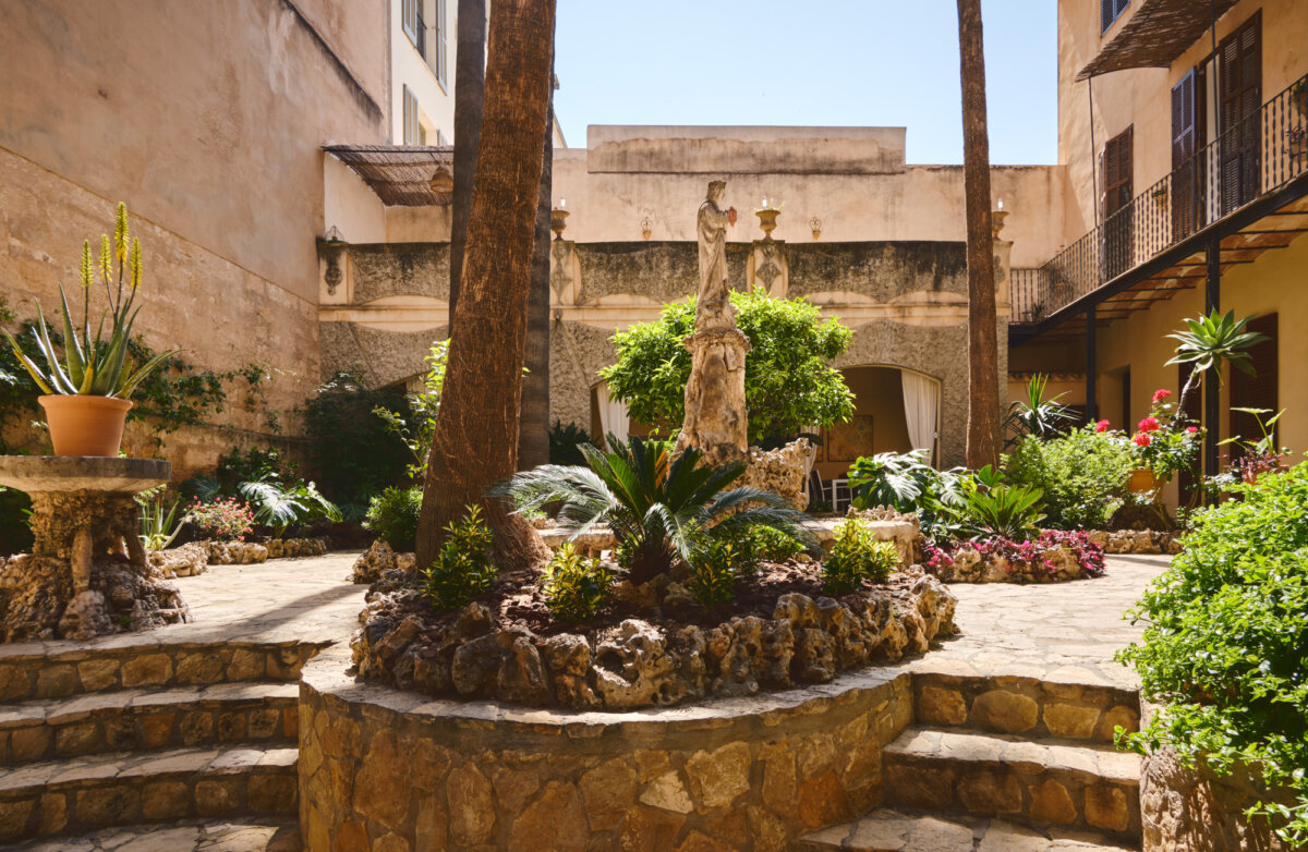 Historischer Innenhof mit Statuen, Palmen und üppigen Pflanzen in mediterraner Umgebung, warme Steinmauern umgeben.