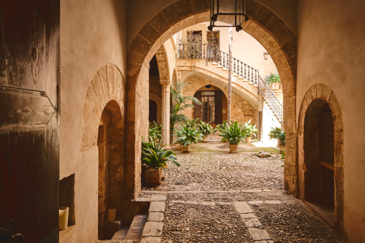 Historischer Innenhof mit gewölbtem Durchgang, Steinboden, Treppe und üppigen Pflanzen in Terrakottatöpfen.