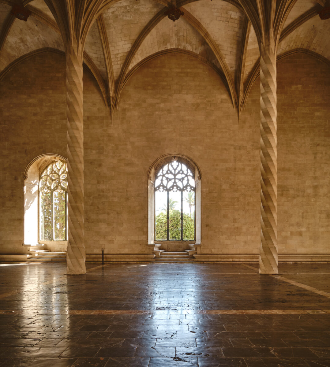 Gotischer Saal mit hohen Gewölben, verzierten Fenstern und Marmorboden. Sonnenlicht fällt durch die reichen Fenster.