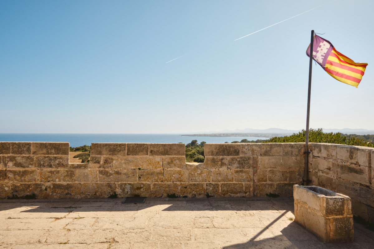 Blick von einer alten Burgmauer auf das Meer bei blauem Himmel, mit einer gelb-roten Flagge im Vordergrund.