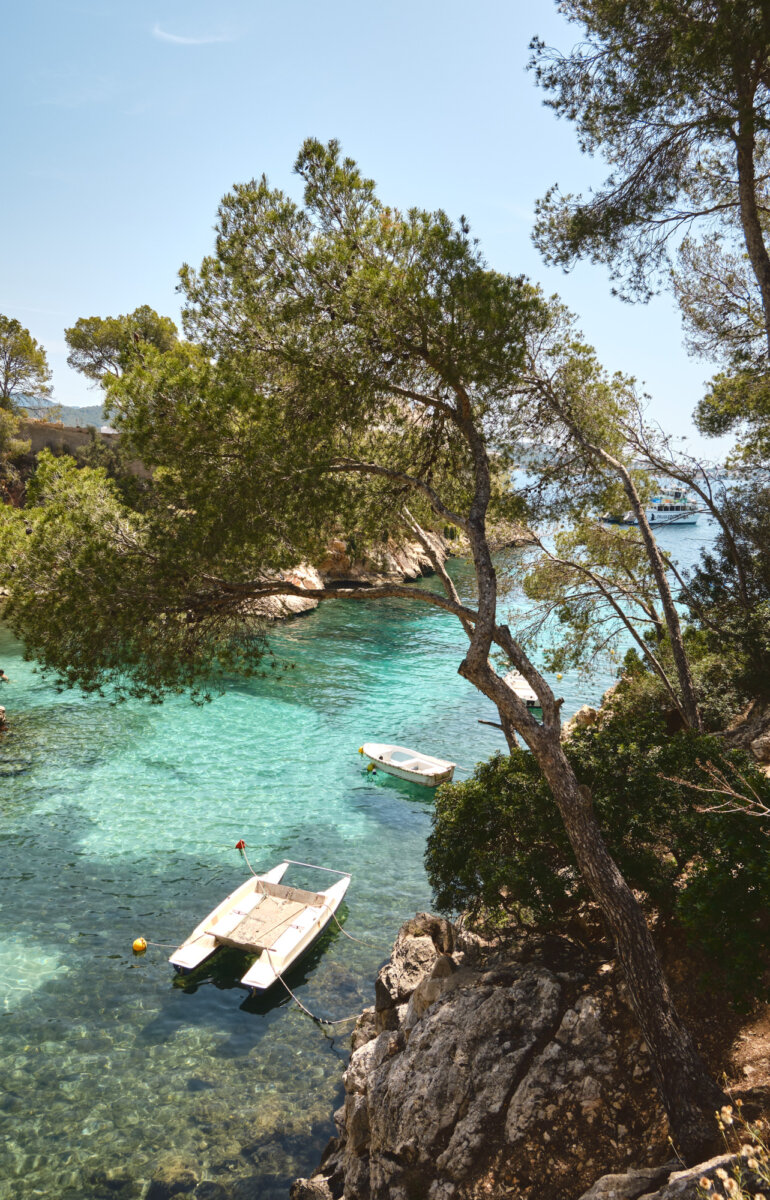 Boote auf türkisfarbenem Meer, umgeben von Bäumen und Felsen in einer ruhigen Bucht unter klarem, blauem Himmel.
