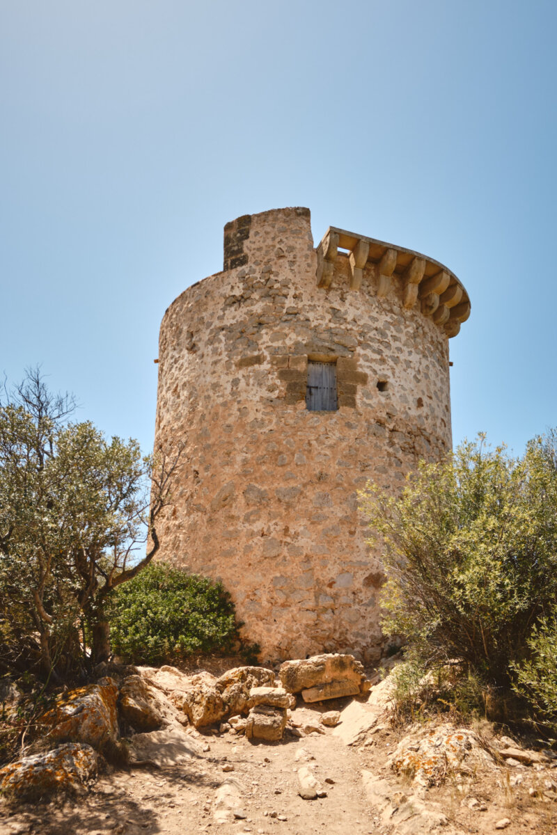 Alter Steinturm an einem sonnigen Tag, umgeben von Büschen und blauem Himmel.