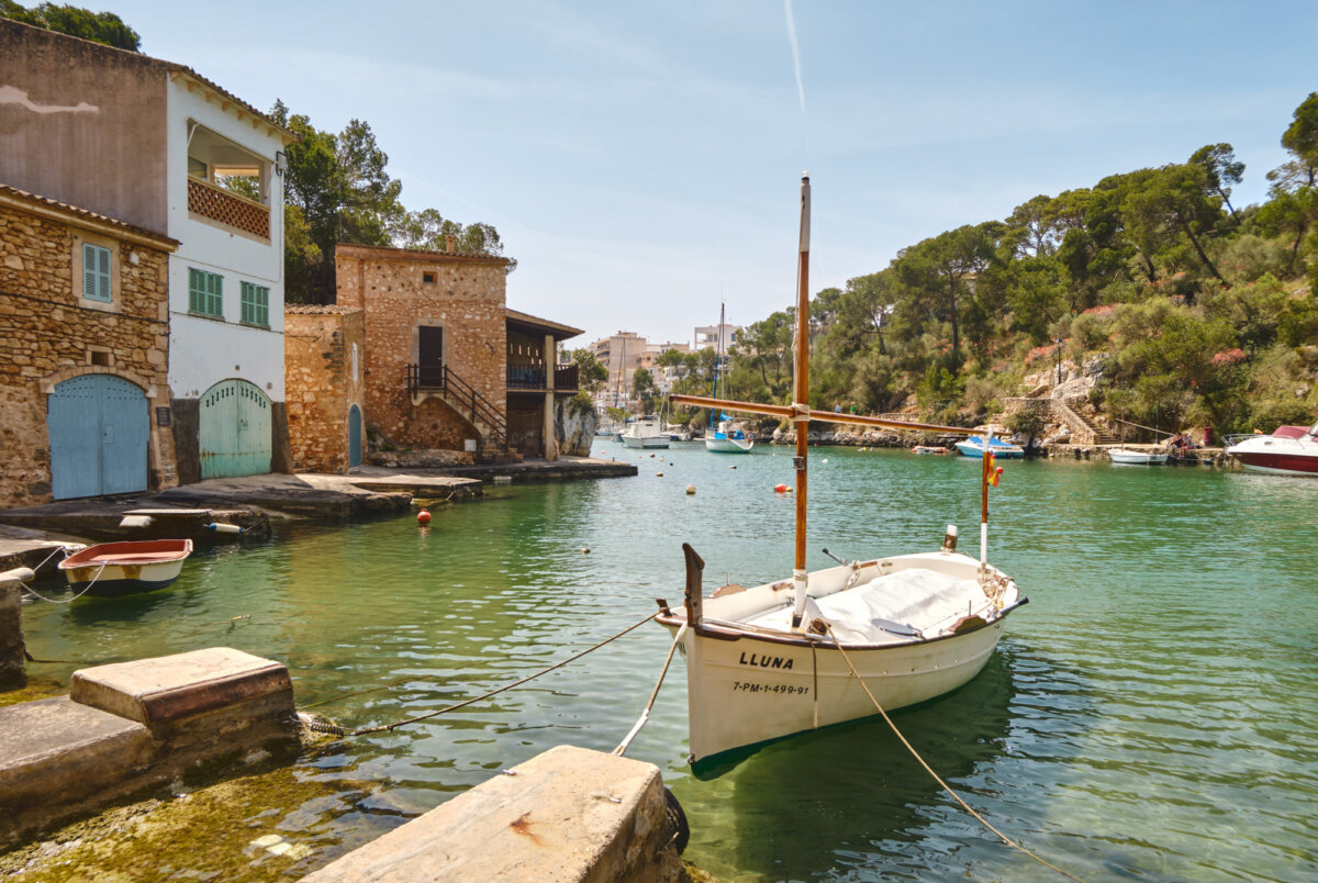Kleines Segelboot in einer malerischen Bucht mit historischen Gebäuden und grünem Wasser bei sonnigem Wetter.