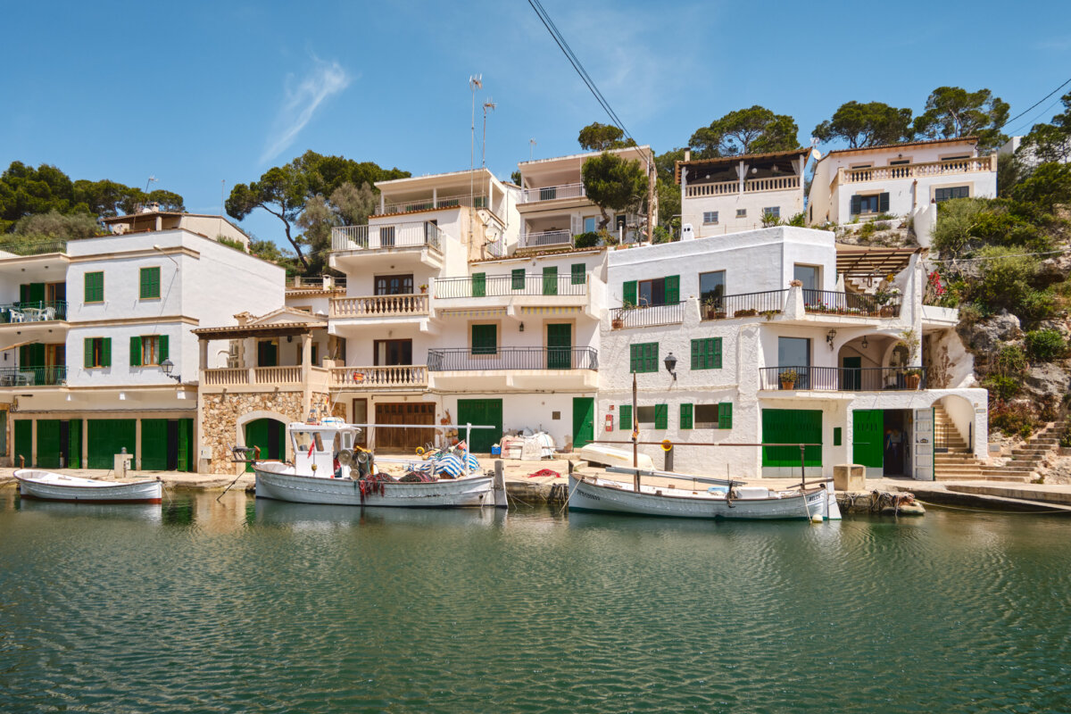 Weiße Hafenhäuser mit grünen Fensterläden am Wasser, Boote im Vordergrund, blauer Himmel im Hintergrund.