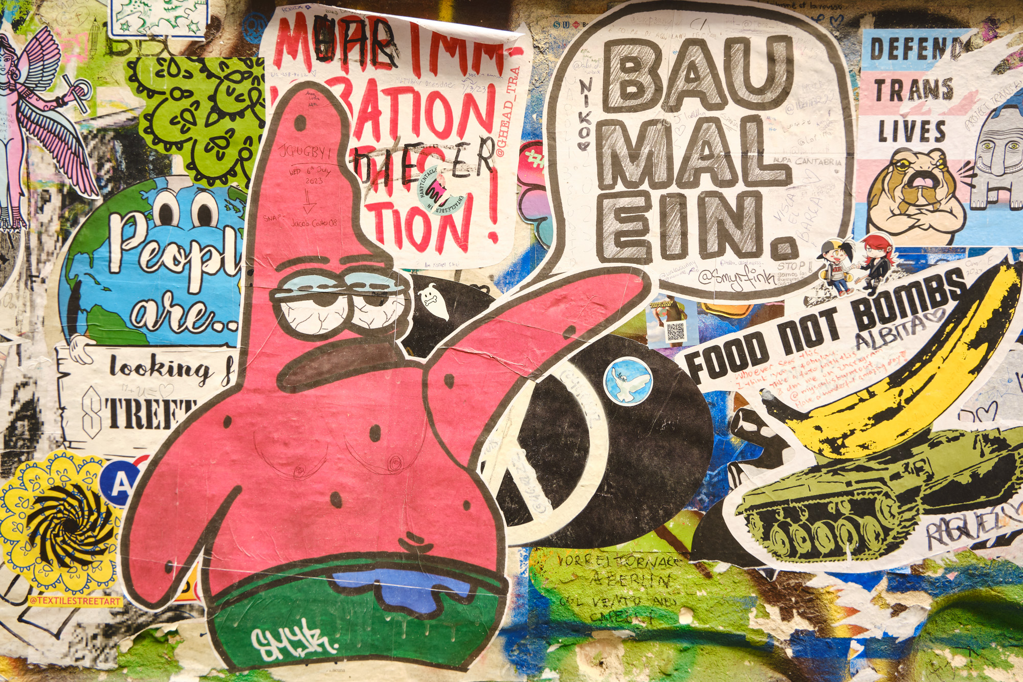 Wand mit bunten Graffitis und Aufklebern, darunter ein rosa Seestern und Texte wie „Baumalein“ und „Food Not Bombs“.