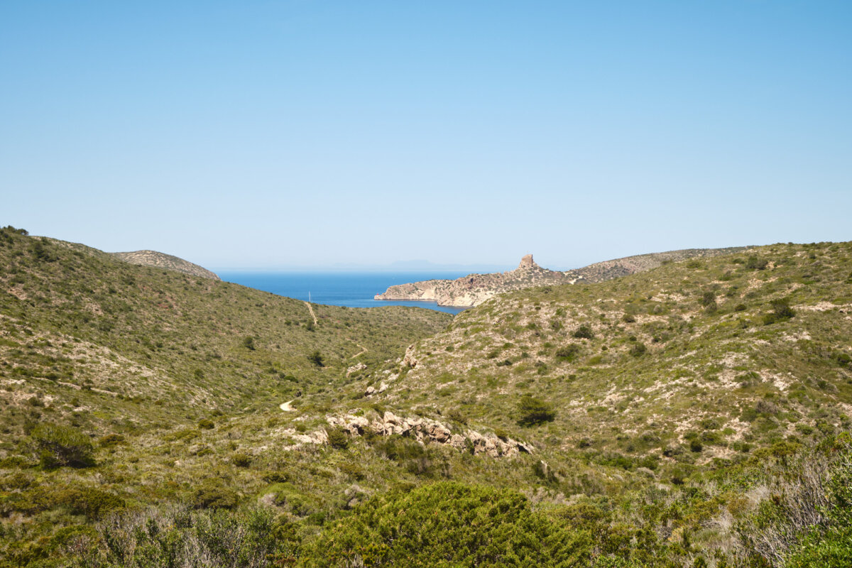 Weite hügelige Landschaft mit grünem Bewuchs und einem entfernten Turm an der Meeresküste unter blauem Himmel.