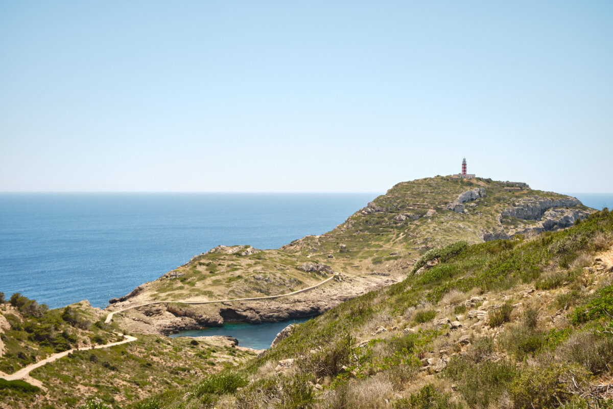 Küstenlandschaft mit einem Leuchtturm auf einem Hügel, umgeben von blauem Meer und grünem Buschwerk unter klarem Himmel.